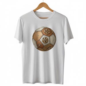 May include: White t-shirt featuring a steampunk-style soccer ball design. The ball is composed of bronze, silver, and gold panels, with gear and rivet details. The shirt is hanging on a wooden hanger.