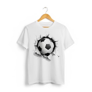 May include: White t-shirt featuring a graphic of a soccer ball bursting through a torn surface. The soccer ball is black and white, with a detailed design. The shirt is hanging on a wooden hanger.