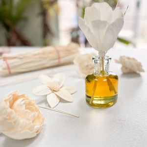 May include: A clear glass bottle with a yellow liquid and a white flower diffuser stick. The bottle is sitting on a white surface with other white flower decorations.