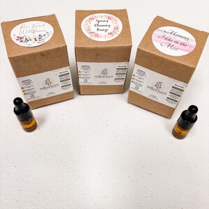 May include: Three brown cardboard boxes with labels featuring floral designs and text. The labels read "Raw Wildflowers", "Spring Cleaning Breeze", and "Morning Hike in the Mist". Each box has a small bottle of essential oil next to it.