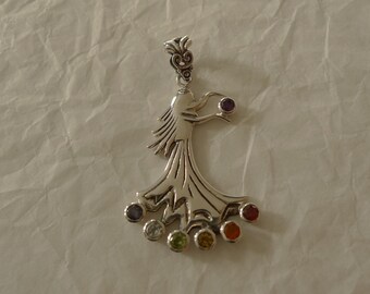 May include: Silver pendant featuring a stylized female figure holding a purple gemstone. The figure is surrounded by seven gemstones in various colors, including red, orange, yellow, green, blue, and white.