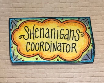 May include: A blue and yellow sign with a cloud shape and the text "Shenanigans Coordinator" in black lettering.