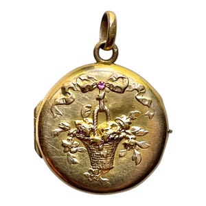 Puede incluir: Un colgante de relicario redondo de metal dorado con un diseño en relieve de una cesta llena de flores y un lazo en la parte superior. El centro del lazo y la cesta de flores están decorados con pequeñas piedras incrustadas de color rosa.