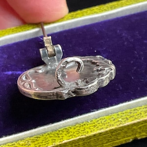 May include: A silver brooch featuring an oval shape with an ornate, embossed border and a central, smooth, light-colored inlay. The back of the brooch displays a metal pin mechanism with a C-shaped clasp and a hinged pin stem for secure attachment.
