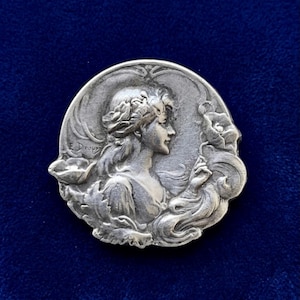 May include: A circular silver-toned metal brooch featuring a relief sculpture of a person in profile with flowing hair and floral elements. The name E. Dropsy is embossed on the left side of the design, which depicts a person holding a flower.
