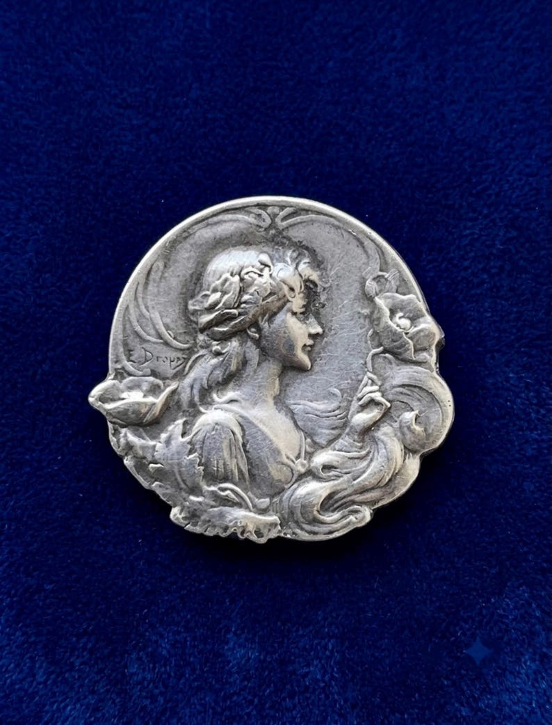 May include: A circular silver-toned metal brooch featuring a relief sculpture of a person in profile with flowing hair and floral elements. The name E. Dropsy is embossed on the left side of the design, which depicts a person holding a flower.