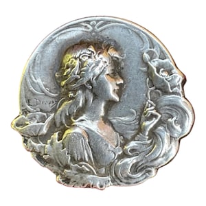 May include: A circular silver-toned metal brooch featuring a relief sculpture of a person in profile, surrounded by stylized floral elements and leaves. The text E. Dropsy is embossed on the left side of the piece, which has a metallic finish.