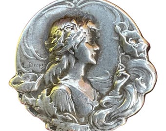 May include: A circular silver-toned metal brooch featuring a relief sculpture of a person in profile, surrounded by stylized floral elements and leaves. The text E. Dropsy is embossed on the left side of the piece, which has a metallic finish.