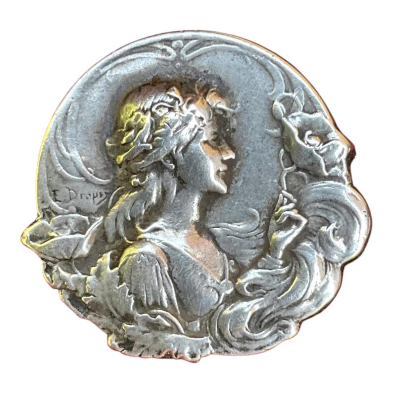 May include: A circular silver-toned metal brooch featuring a relief sculpture of a person in profile, surrounded by stylized floral elements and leaves. The text E. Dropsy is embossed on the left side of the piece, which has a metallic finish.