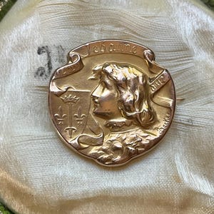 May include: A round gold-toned brooch featuring a relief profile of a person with wavy hair. The text Jehanne is inscribed on a banner above the head. To the left, a shield displays a crown and three fleur-de-lis symbols. The name Becker is visible.