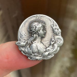 May include: A circular silver-toned metal brooch featuring a relief profile of a person with flowing hair surrounded by floral motifs. The name E. Dropy is embossed on the left side of the piece. The design includes intricate details of leaves and petals.
