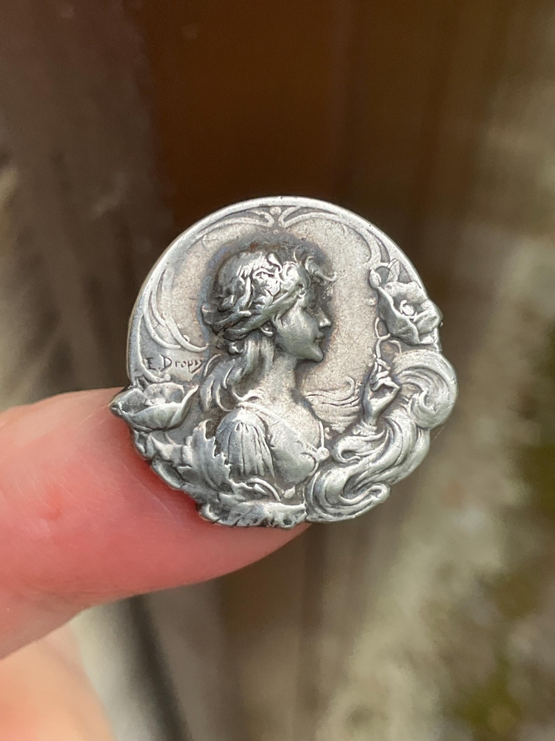 May include: A circular silver-toned metal brooch featuring a relief profile of a person with flowing hair surrounded by floral motifs. The name E. Dropy is embossed on the left side of the piece. The design includes intricate details of leaves and petals.
