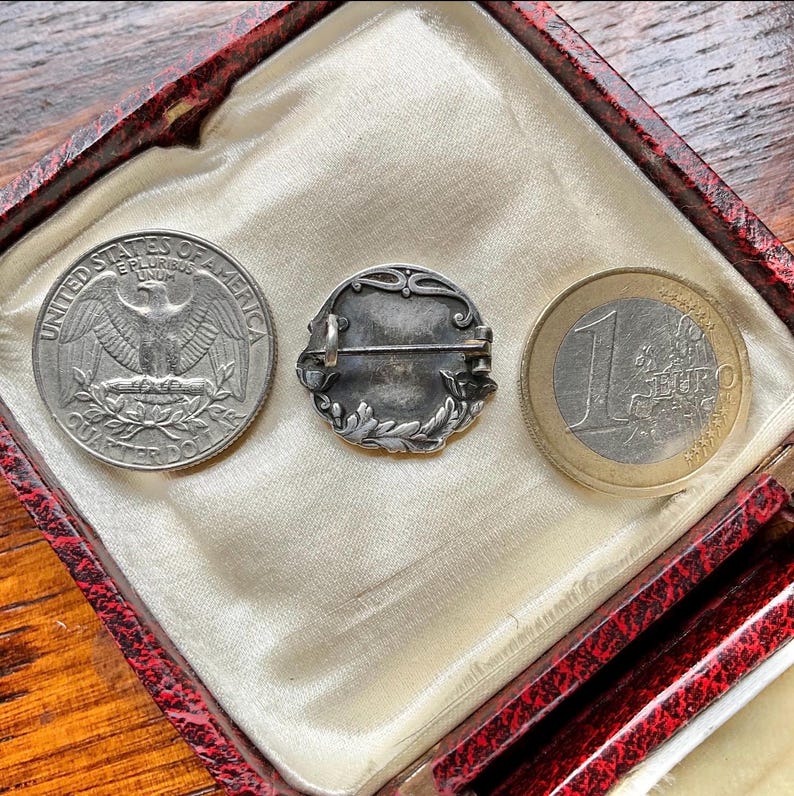 May include: A circular silver-toned metal brooch featuring an ornate leaf-patterned border and a horizontal pin mechanism on the back, displayed between a United States quarter dollar and a one-euro coin for scale reference.