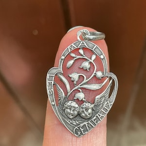 May include: A silver-toned metal pendant shaped like a heart, featuring two cherub faces at the bottom and a lily of the valley floral motif above. The text "LES GEMEAUX" is embossed along the curved border of the frame, with a small metal loop at the top.