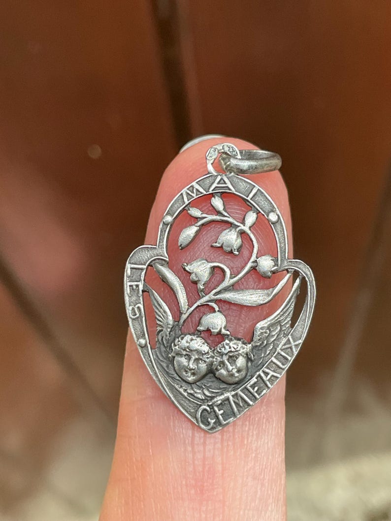 May include: A silver-toned metal pendant shaped like a heart, featuring two cherub faces at the bottom and a lily of the valley floral motif above. The text "LES GEMEAUX" is embossed along the curved border of the frame, with a small metal loop at the top.