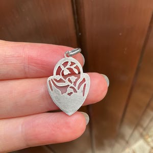 May include: A silver-toned metal pendant shaped like a heart with a pointed base. The pendant features an intricate cutout design depicting floral stems with small blossoms within the upper arched section and a solid matte finish below.