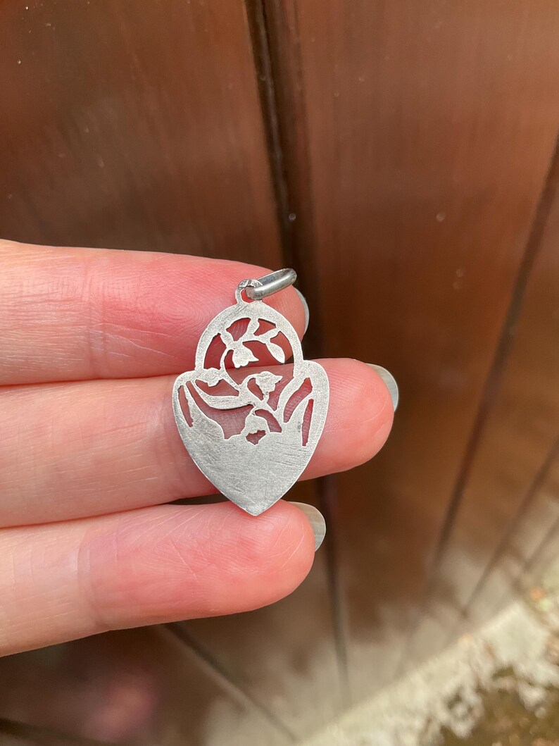 May include: A silver-toned metal pendant shaped like a heart with a pointed base. The pendant features an intricate cutout design depicting floral stems with small blossoms within the upper arched section and a solid matte finish below.