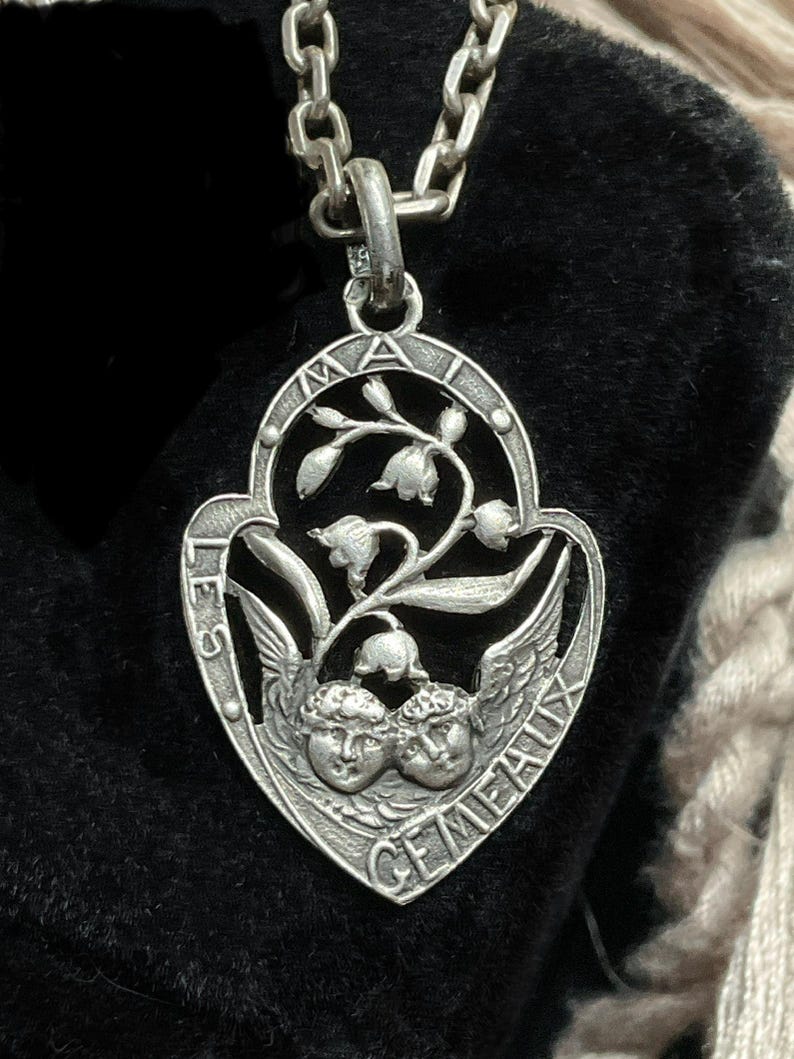 May include: A silver-toned metal pendant on a matching chain, featuring an openwork design with two cherub faces at the bottom, lily of the valley flowers above, and the text LES GEMEAUX MAI engraved along the curved border.