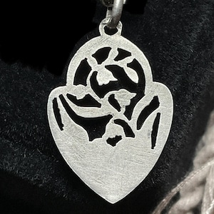 May include: A silver-toned pendant shaped like a heart with a pointed base, featuring an intricate cutout design of floral elements. The pendant is suspended from a matching silver-toned metal chain link necklace.