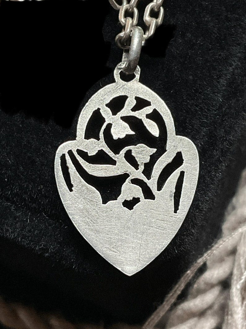 May include: A silver-toned pendant shaped like a heart with a pointed base, featuring an intricate cutout design of floral elements. The pendant is suspended from a matching silver-toned metal chain link necklace.