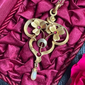 May include: A gold pendant necklace featuring two stylized floral motifs with small red gemstones at their centers, a central silver-toned crescent element, and a single teardrop-shaped white pearl dangling from the bottom of the frame.