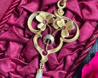 May include: A gold pendant necklace featuring two stylized floral motifs with small red gemstones at their centers, a central silver-toned crescent element, and a single teardrop-shaped white pearl dangling from the bottom of the frame.