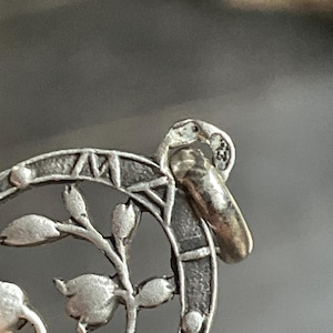 May include: A silver-toned metal pendant featuring a circular frame with engraved Roman numerals and a central floral motif. A small, thick jump ring is attached to the top, displaying two distinct, small stamped hallmarks.