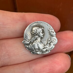 May include: A circular silver-toned metal brooch featuring a relief portrait of a person in profile, surrounded by flowing floral motifs and leaves. The name E. Dropsy is engraved on the left side of the piece, which has a polished finish.