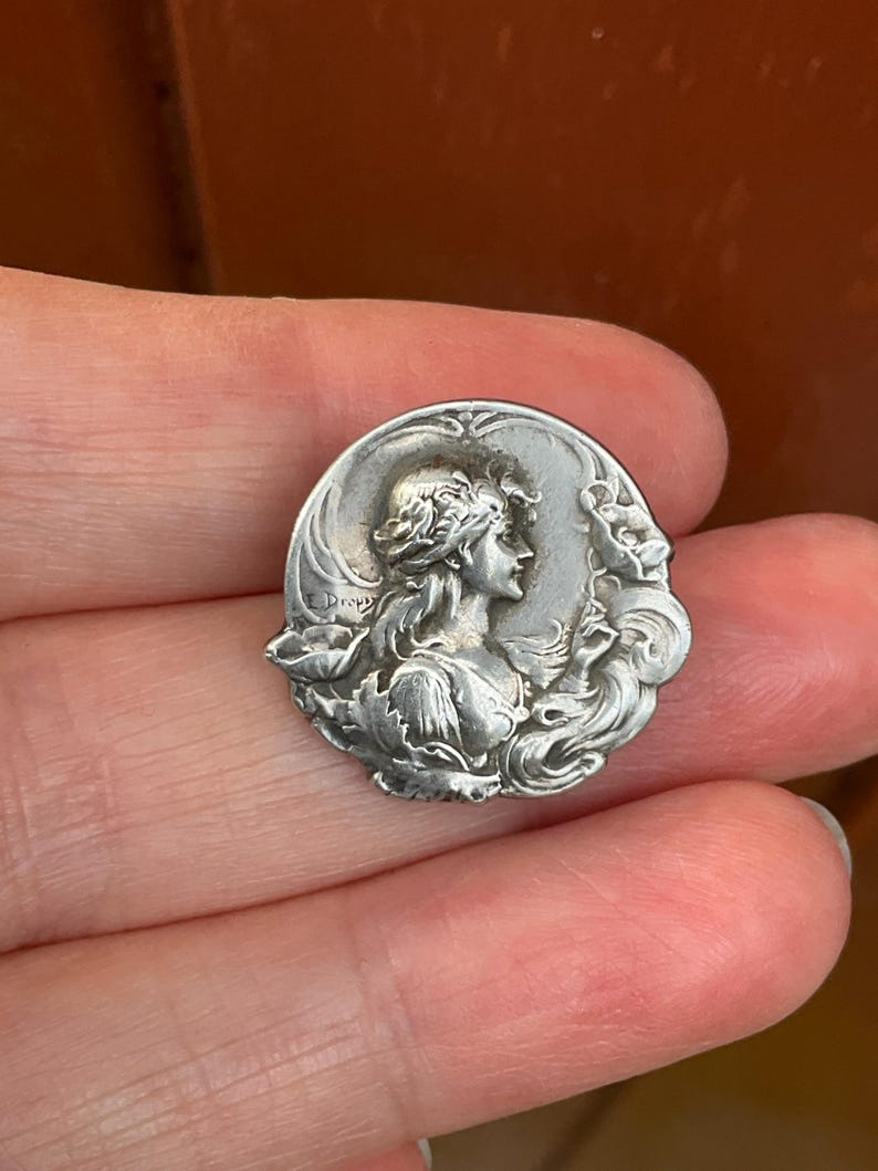 May include: A circular silver-toned metal brooch featuring a relief portrait of a person in profile, surrounded by flowing floral motifs and leaves. The name E. Dropsy is engraved on the left side of the piece, which has a polished finish.