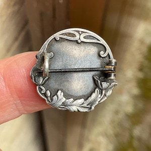May include: A circular silver-toned metal brooch featuring an embossed floral border with leaves and flower buds. The back includes a horizontal pin mechanism with a hinge and a catch, showing a tarnished metallic finish and intricate relief details.