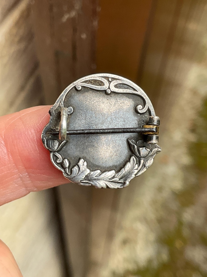 May include: A circular silver-toned metal brooch featuring an embossed floral border with leaves and flower buds. The back includes a horizontal pin mechanism with a hinge and a catch, showing a tarnished metallic finish and intricate relief details.