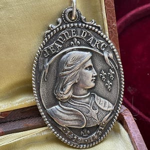 May include: An oval silver-toned metal pendant featuring a relief profile portrait of Jeanne d'Arc in armor. The text 'JEANNE D'ARC' is inscribed at the top, with '1412' and '1431' at the bottom, accented by three fleur-de-lis symbols on the side.