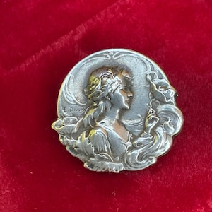 May include: A circular silver-toned metal brooch featuring a relief portrait of a person in profile with flowing hair, surrounded by stylized floral elements. The name E. Dropsy is embossed on the left side of the metallic surface.