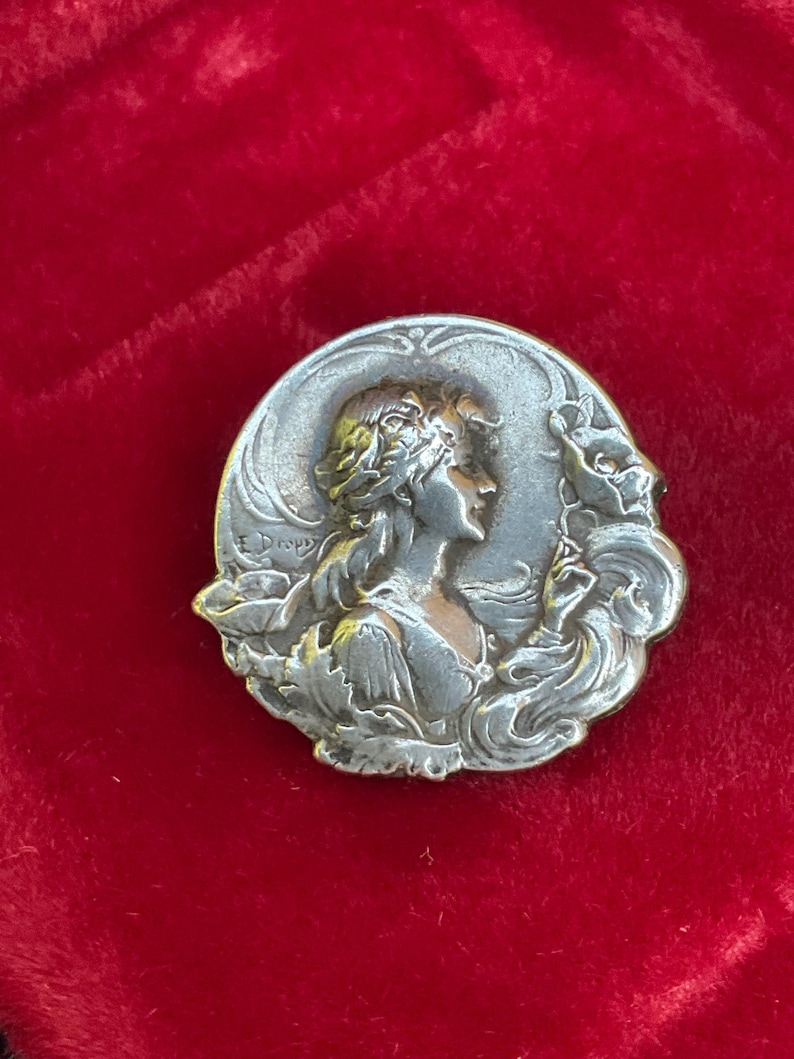 May include: A circular silver-toned metal brooch featuring a relief portrait of a person in profile with flowing hair, surrounded by stylized floral elements. The name E. Dropsy is embossed on the left side of the metallic surface.