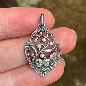 May include: A silver-toned metal pendant featuring an openwork design of lily of the valley flowers and two cherub faces at the base. The text "LES GEMEAUX" and "MAI" is engraved along the curved border of the shield-shaped charm.