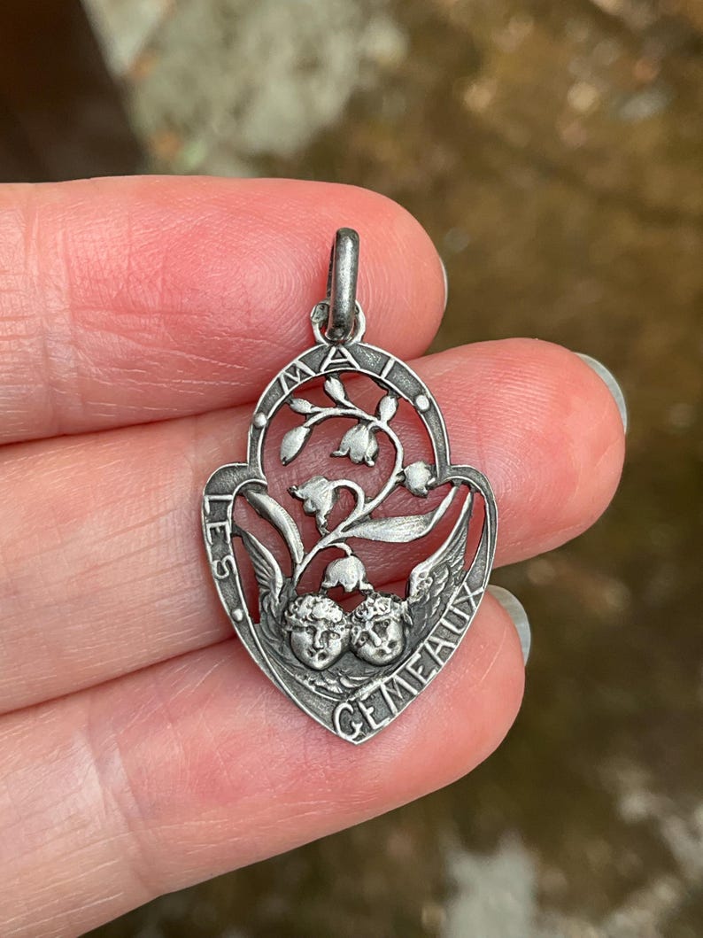 May include: A silver-toned metal pendant featuring an openwork design of lily of the valley flowers and two cherub faces at the base. The text "LES GEMEAUX" and "MAI" is engraved along the curved border of the shield-shaped charm.