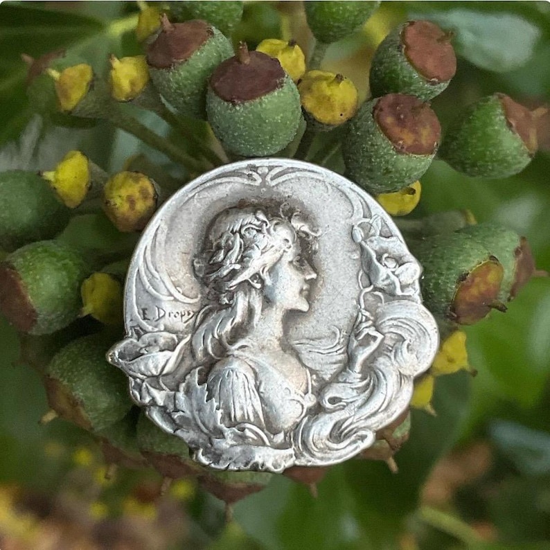 May include: A circular silver-toned metal medallion featuring a relief profile of a person with flowing hair, surrounded by floral motifs and the engraved text E. Dropsy. The piece has a decorative border and a polished, reflective finish.