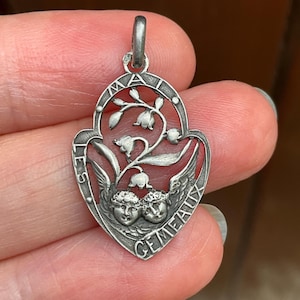 May include: A silver-toned metal pendant featuring an openwork design of lily of the valley flowers above two cherub faces. The border is inscribed with the text "LES MAI" and "GEMEAUX" along the curved edges, with a small bail at the top.