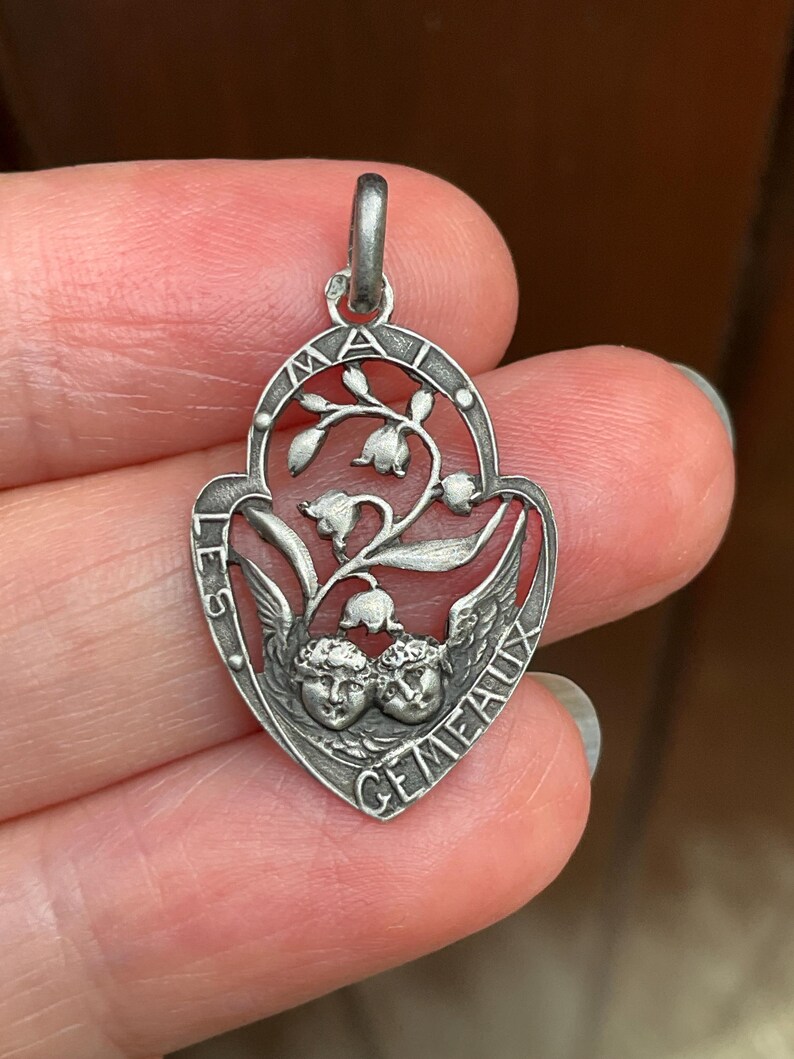May include: A silver-toned metal pendant featuring an openwork design of lily of the valley flowers above two cherub faces. The border is inscribed with the text "LES MAI" and "GEMEAUX" along the curved edges, with a small bail at the top.