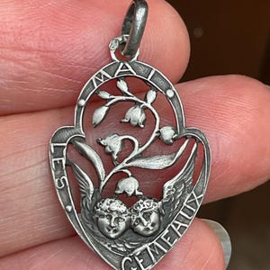 May include: A silver-toned metal pendant featuring an openwork design of two cherub faces at the bottom and lily of the valley flowers above. The frame is inscribed with the text LES GEMEAUX and MAI, with a small loop at the top for attachment.