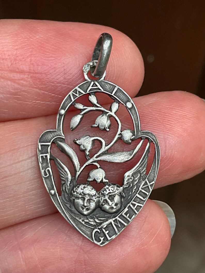 May include: A silver-toned metal pendant featuring an openwork design of two cherub faces at the bottom and lily of the valley flowers above. The frame is inscribed with the text LES GEMEAUX and MAI, with a small loop at the top for attachment.