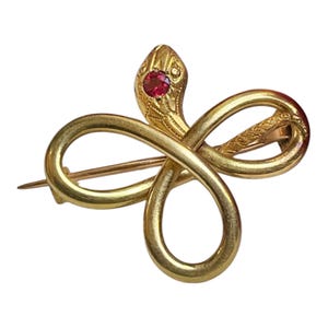 Peut inclure: Une broche dorée en forme de serpent enroulé formant une boucle à l'infini. La tête du serpent présente une petite pierre précieuse ronde d'un rouge vif sertie au centre. Le corps du serpent a une finition lisse et polie avec des détails texturés.