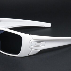 May include: White sunglasses with dark lenses, displayed on a black backdrop. The sunglasses feature a contemporary, sporty design with a bold frame and arms. The Oakley logo is visible on the temple.