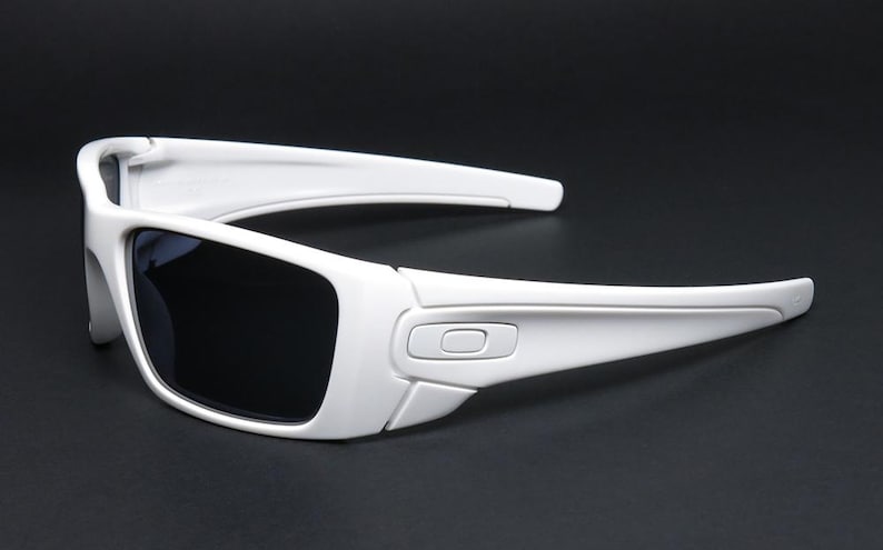 May include: White sunglasses with dark lenses, displayed on a black backdrop. The sunglasses feature a contemporary, sporty design with a bold frame and arms. The Oakley logo is visible on the temple.