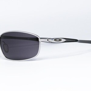 May include: Silver-framed sunglasses with dark gray lenses. The sunglasses have a sleek, modern design with a curved frame and black temples. The Oakley logo is visible on the temple. The sunglasses are set against a white background.