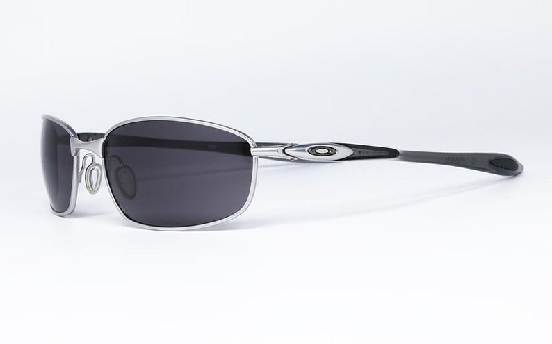 May include: Silver-framed sunglasses with dark gray lenses. The sunglasses have a sleek, modern design with a curved frame and black temples. The Oakley logo is visible on the temple. The sunglasses are set against a white background.