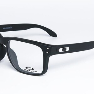 May include: Black rectangular eyeglasses with clear lenses. The matte black frames have the Oakley logo on the temples and lens. The model name "HOLBROOK RX" is printed on the top of the frame.