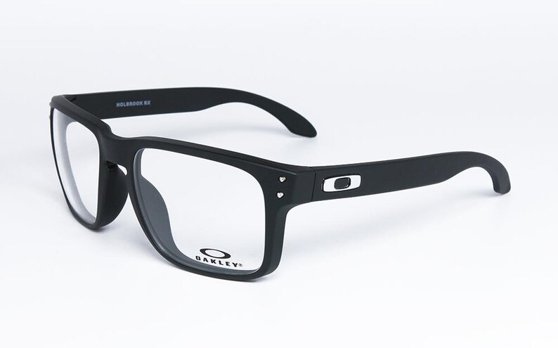 May include: Black rectangular eyeglasses with clear lenses. The matte black frames have the Oakley logo on the temples and lens. The model name "HOLBROOK RX" is printed on the top of the frame.