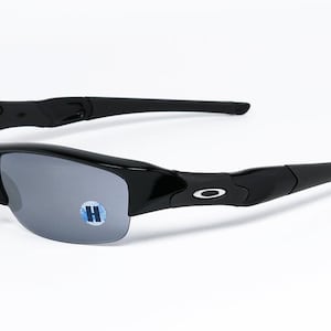 K&ouml;nnte beinhalten: Schwarze Sonnenbrille mit grauen Gl&auml;sern. Die Sonnenbrille hat ein halb rahmenloses Design und einen geschwungenen Rahmen. Das Oakley-Logo ist auf den B&uuml;geln sichtbar. Ein kleiner blau-wei&szlig;er Aufkleber mit dem Buchstaben "H" befindet sich auf dem Glas.