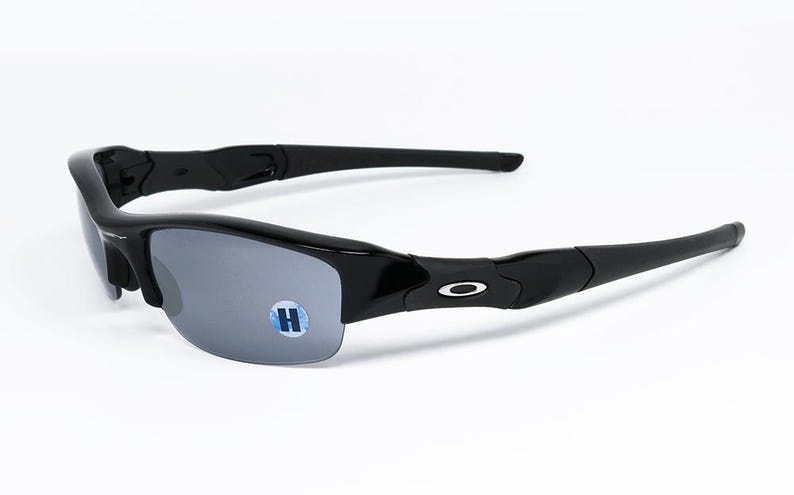 K&ouml;nnte beinhalten: Schwarze Sonnenbrille mit grauen Gl&auml;sern. Die Sonnenbrille hat ein halb rahmenloses Design und einen geschwungenen Rahmen. Das Oakley-Logo ist auf den B&uuml;geln sichtbar. Ein kleiner blau-wei&szlig;er Aufkleber mit dem Buchstaben "H" befindet sich auf dem Glas.